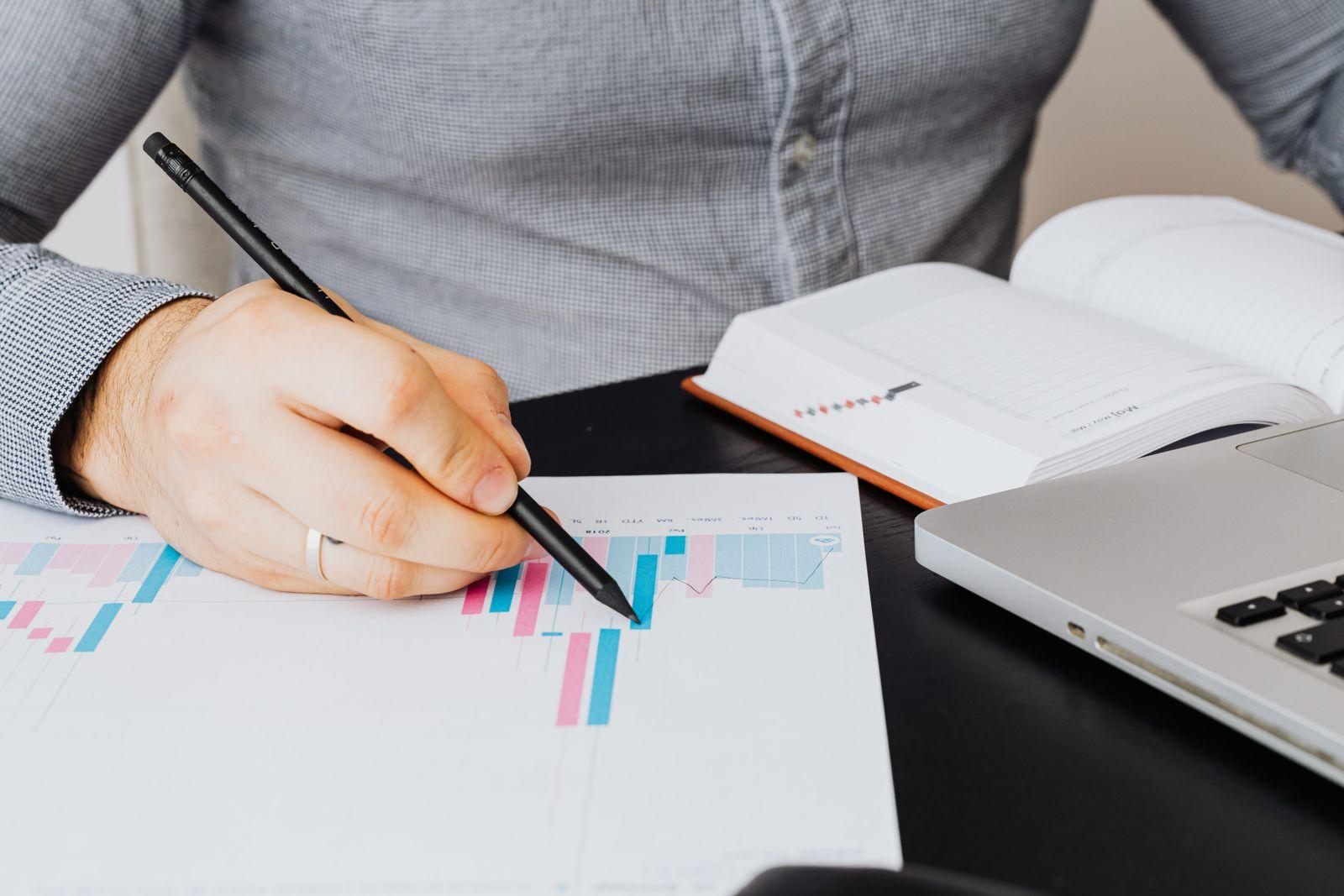 Hand with a pen marking a printed bar chart next to a laptop