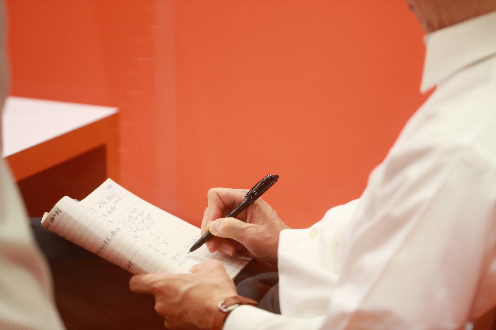 Person in a white shirt writing notes in a spiral notepad during a meeting
