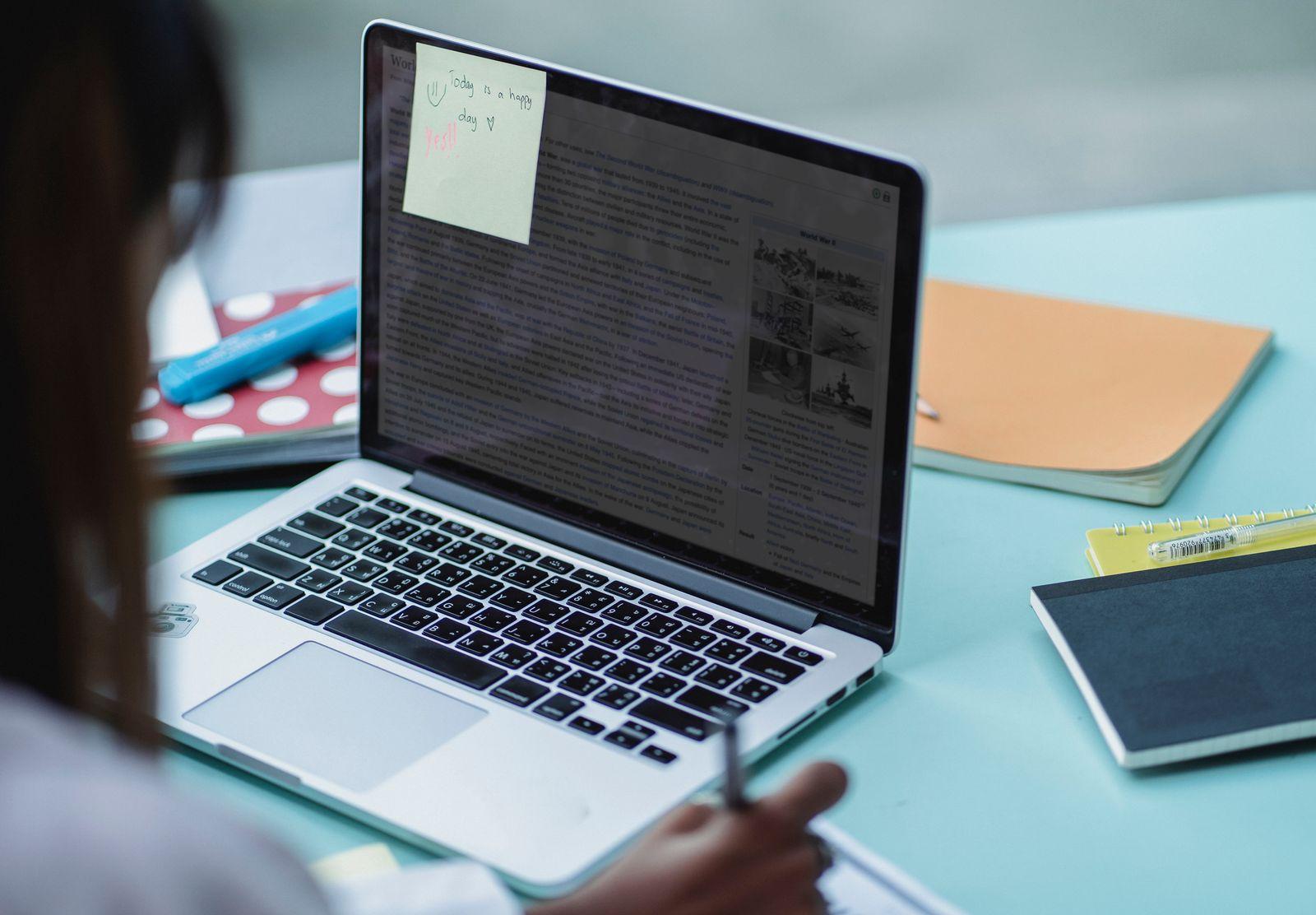 Student writing notes beside a laptop with a sticky note on the screen