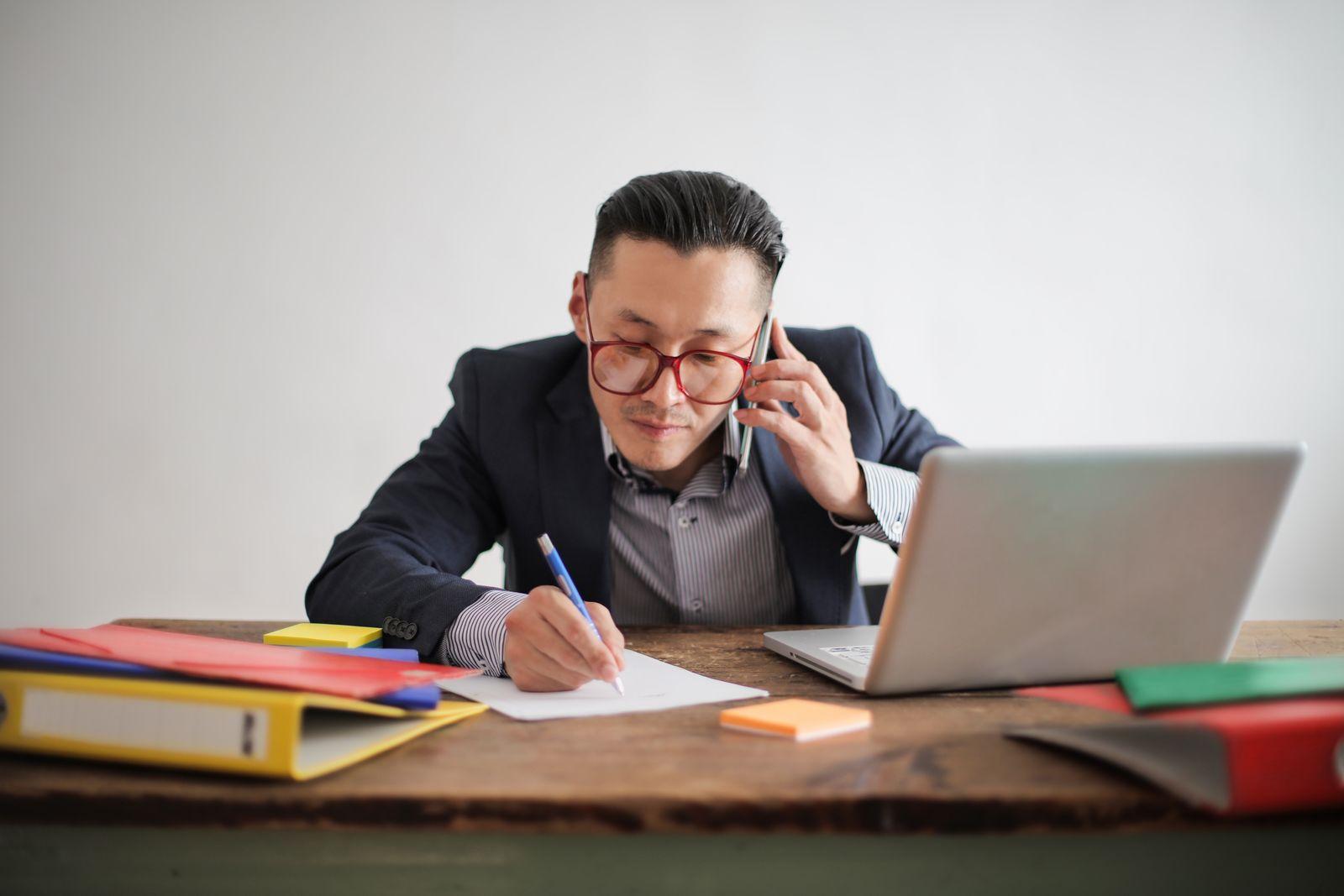 Man on a phone call taking notes at a desk with a laptop