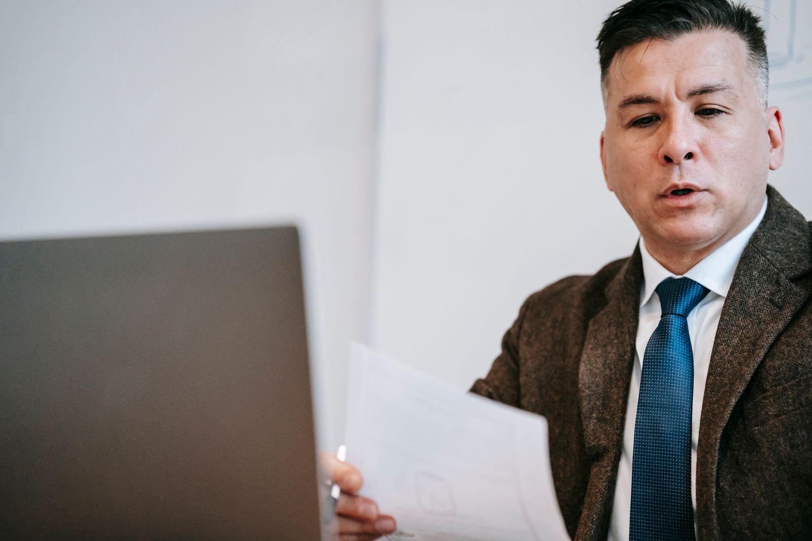 Presenter in a suit speaking toward a laptop while holding notes