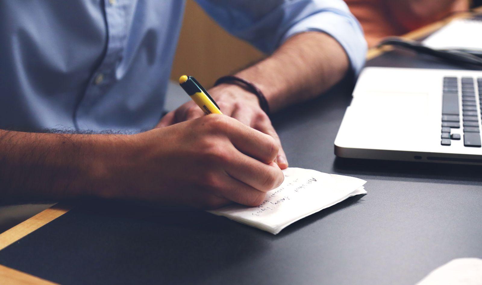 Person writing on a notepad next to an open laptop