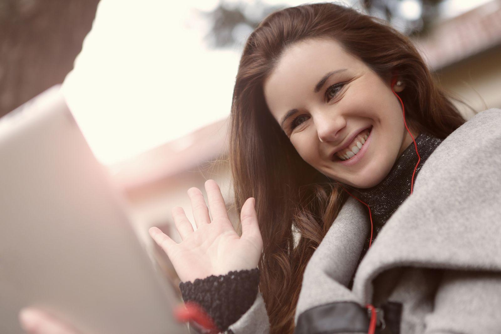 Smiling woman with earphones waving at a laptop during a video call